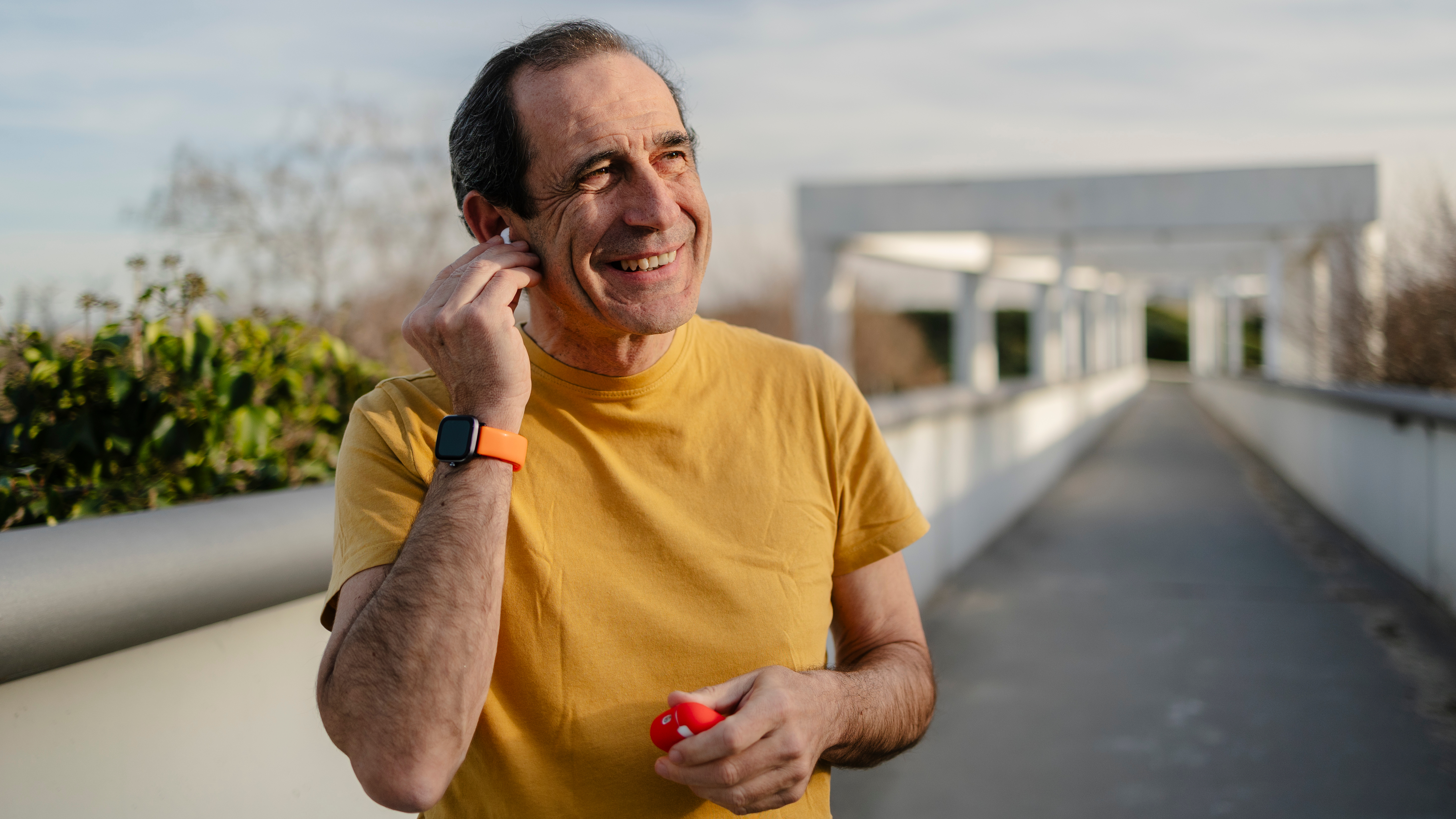 A smiling man stands outside wearing a smartwatch and putting in earbuds. Behind him we see a long bridge and some greenery.