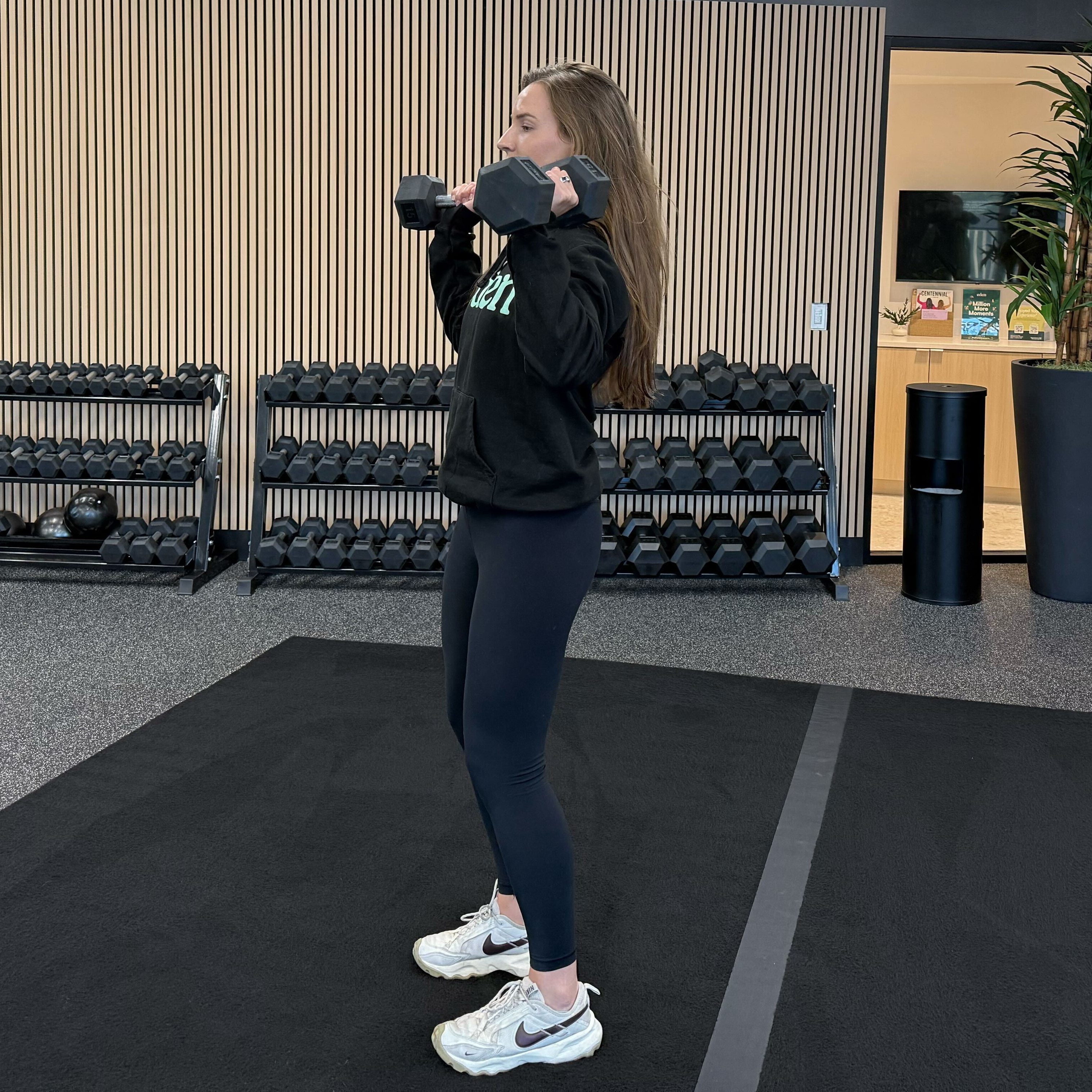 Felicia Hernandez, a personal trainer for Eden Health Club, performs a dumbbell shoulder press in a fitness studio. She is standing with feet shoulder-width apart, dumbbells held at shoulder height.