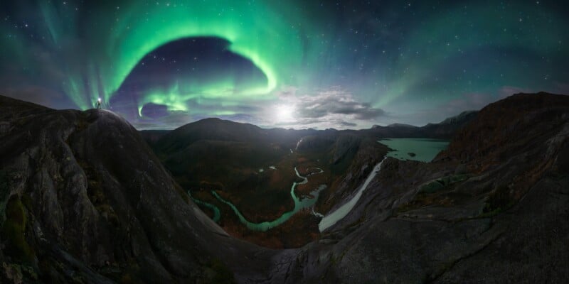 A vibrant green aurora borealis lights up the night sky over a rugged mountain landscape with winding rivers, rocky terrain, and distant lakes under a star-filled sky.