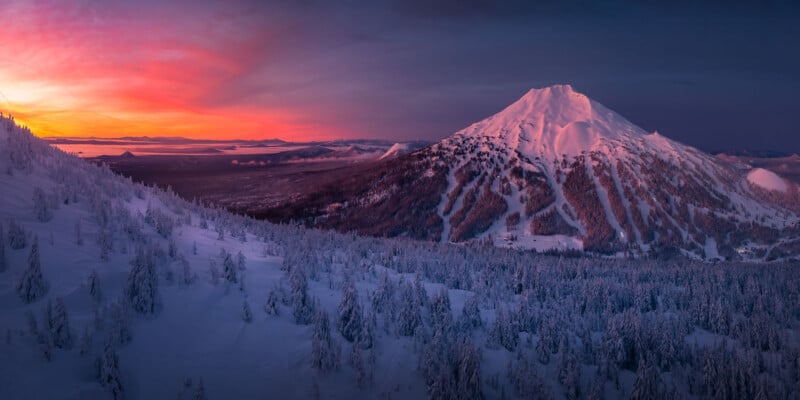 Snow-covered mountain and forest at sunset, with vibrant pink, orange, and purple clouds in the sky and the sun low on the horizon, casting a warm glow over the wintry landscape.