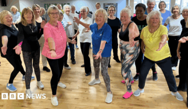 A large group of cheerful people wearing trainers and sports clothing surround dance instructor Maureen Conroy. They are standing in a large gym which has a polished pine floor.