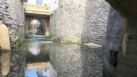 Lyme Regis Town Council The River Lim viewed from beneath a stone bridge. The view is far below street level and the river is running through a deep channel with high stone walls either side.