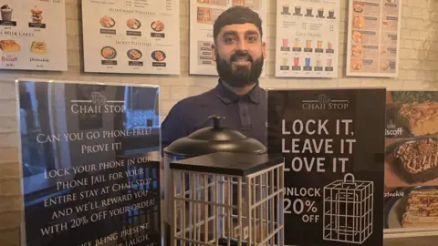 Usman Hussain Usman Hussain, who has short dark hair and a beard and is wearing a navy polo shirt, stands in a cafe behind the "phone jail" - a white wire box between two signs that say "lock it, leave it, love it"