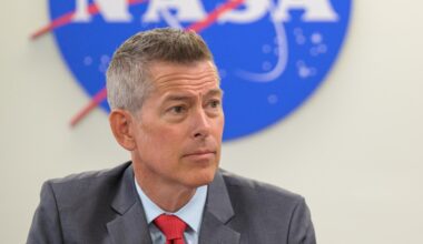 A man in a suit sit looks to the right in front of the bottom half of a nasa meatball logo.