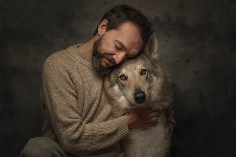 A man with a beard wearing a beige sweater gently hugs and leans his head against a large, wolf-like dog, both appearing calm and close against a dark, textured background.