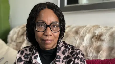 BBC Paula Robinson sat on a settee wearing a leopard print jacket. She has long black hair and is wearing wide, black-rimmed glasses. 
