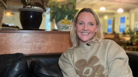Kate is sitting on a black leather couch in a cozy indoor setting with warm lighting. Behind her, there are potted plants on a wooden partition and hanging lights illuminating the space. The background shows a spacious room with tables and chairs