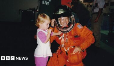 Eileen Collins wearing a blue jacket with a round Nasa patch on one side and a rectangular shuttle patch on the other with her name. She has short brown hair and brown eyes and is smiling as she looks directly to camera. She is in the space gallery at the Science Museum and an out of focus rocket engine can be seen behind her.