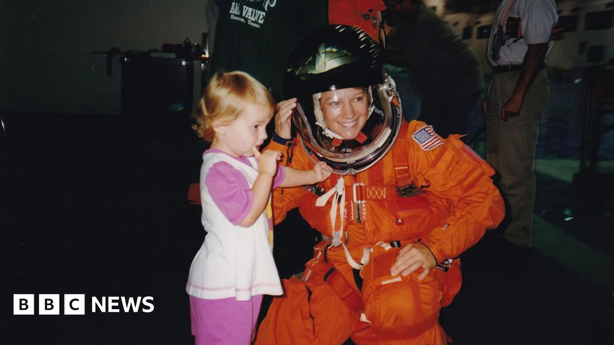 Eileen Collins wearing a blue jacket with a round Nasa patch on one side and a rectangular shuttle patch on the other with her name. She has short brown hair and brown eyes and is smiling as she looks directly to camera. She is in the space gallery at the Science Museum and an out of focus rocket engine can be seen behind her.