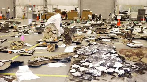 NASA Debris from the Space Shuttle Columbia is laid out on the floor of a large hanger. A man in a white coat is bending down inspecting some of it.  There are tiles and pieces of white foam - some of the material is charred. 