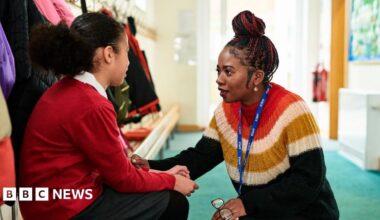 The image shows a woman and a girl in a school cloakroom, with the little girl sat on a bench while the woman crouches in front of her. The woman, a teacher, is wearing a striped jumper while holding a pair of glasses with black frames. The little girl is wearing a red jumper and a grey skirt with black tights. The floor is green and coats of different colours can be seen in the background.