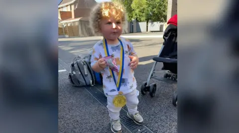 Jay Baggaley Hunter is standing and holding a packet of sweets. He has short curly blonde hair. He has a gold medal with a yellow and blue ribbon strap around his neck.