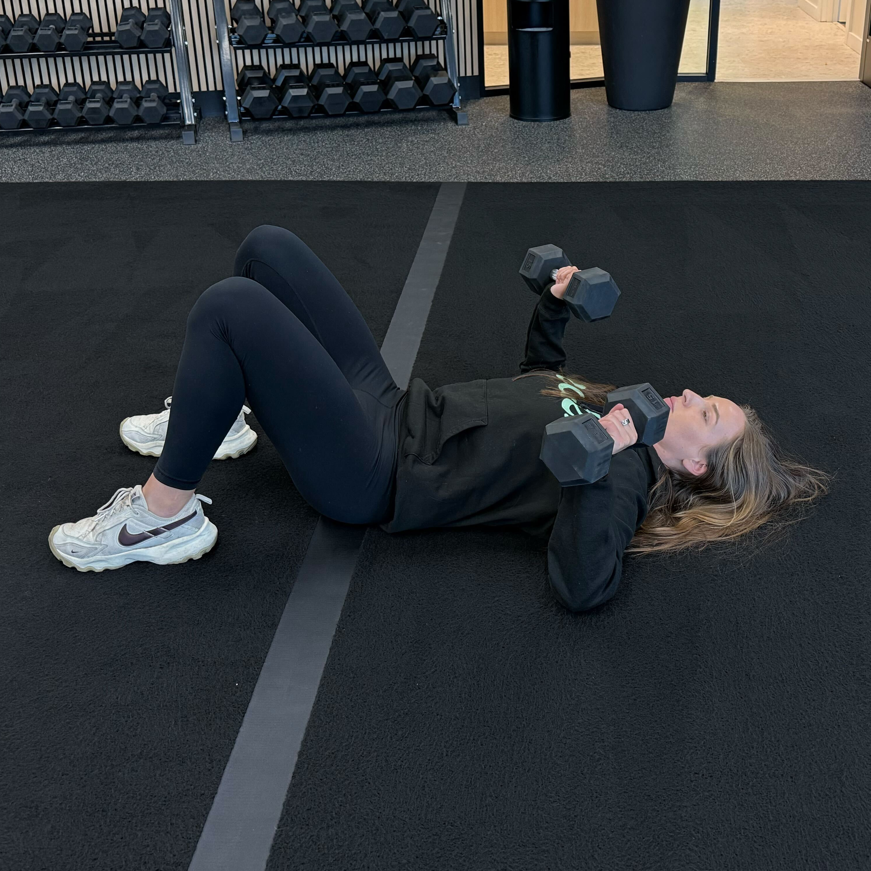 Felicia Hernandez, a personal trainer for Eden Health Club, performs a dumbbell chest press on the floor. She is lying with her knees bent and feet flat on the floor, elbows bent and dumbbells close to her shoulders. 