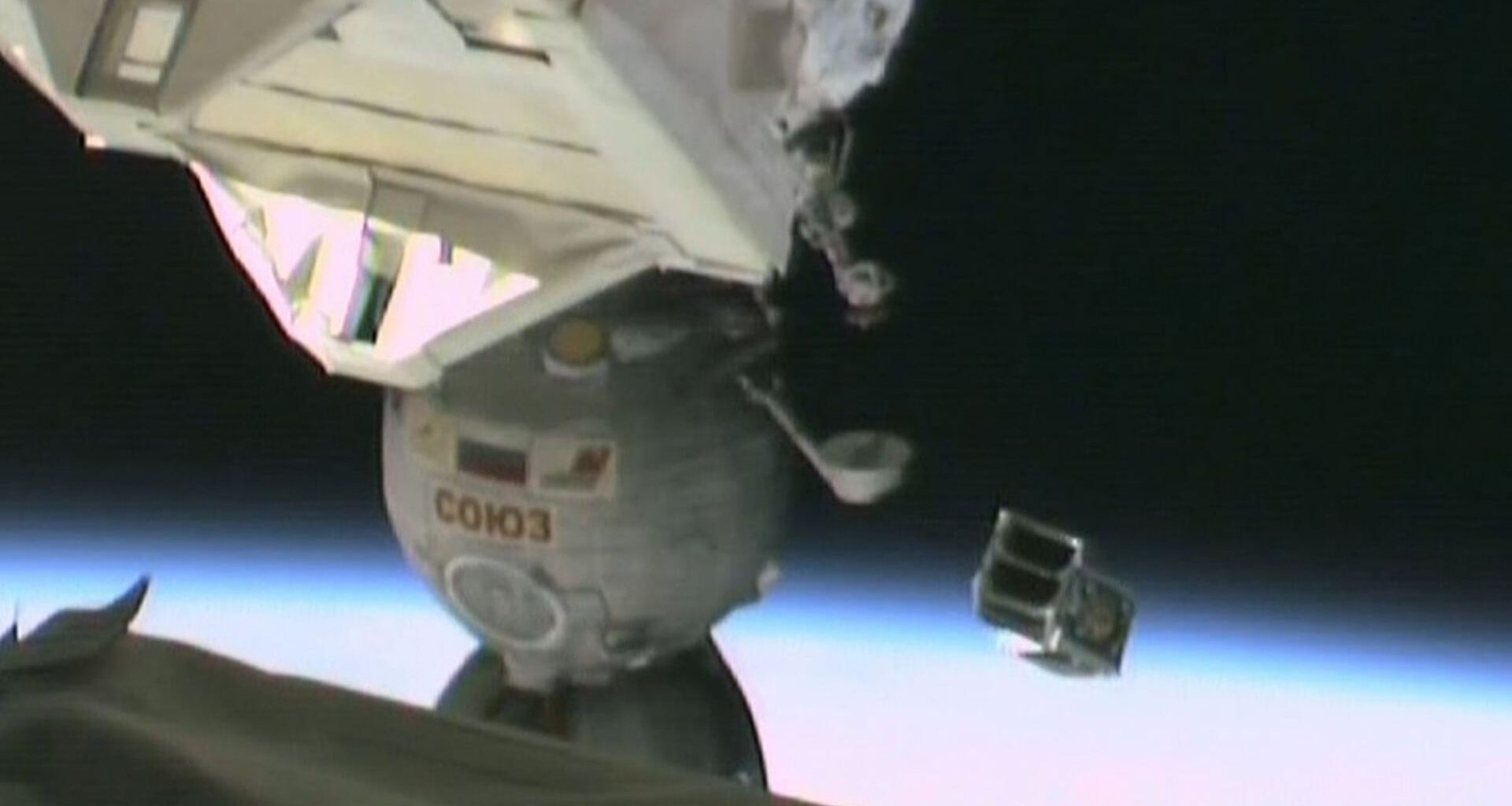 A small metal satellite is launched floating above Earth with part of the International Space Station seen on the left part of the image