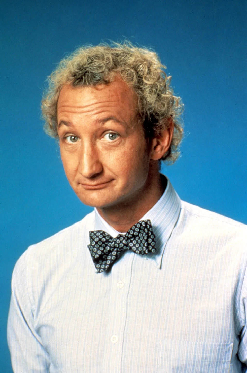 Person with curly hair wearing a patterned bow tie and striped shirt against a plain backdrop