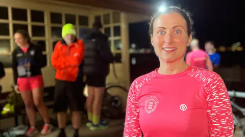 BBC Katie is dressed in a bright pink long-sleeve athletic top with "Jersey Spartan AC" printed on a race bib numbered "2". The setting is outdoors at night with other runners and bicycles in the background near a building