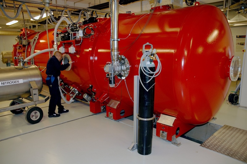 Long red tubular metal tank, with various pipes and other equipment attached to it. A man is looking inside a round opening on the tank.