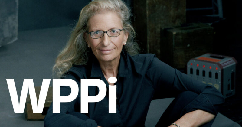 A woman with long, wavy blonde hair and glasses, wearing a black shirt, sits with her arms resting on her knees. Large white letters "WPPI" are overlaid on the image.