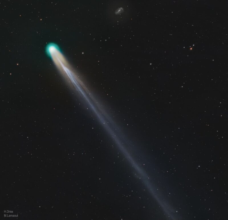 A comet with a green head and intricate tail with a distant spiral galaxy small at the top.