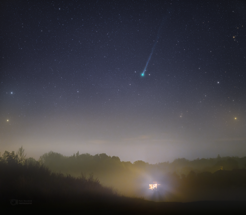 Foggy light at night in the valley with a greenish-blue long-tailed comet above.
