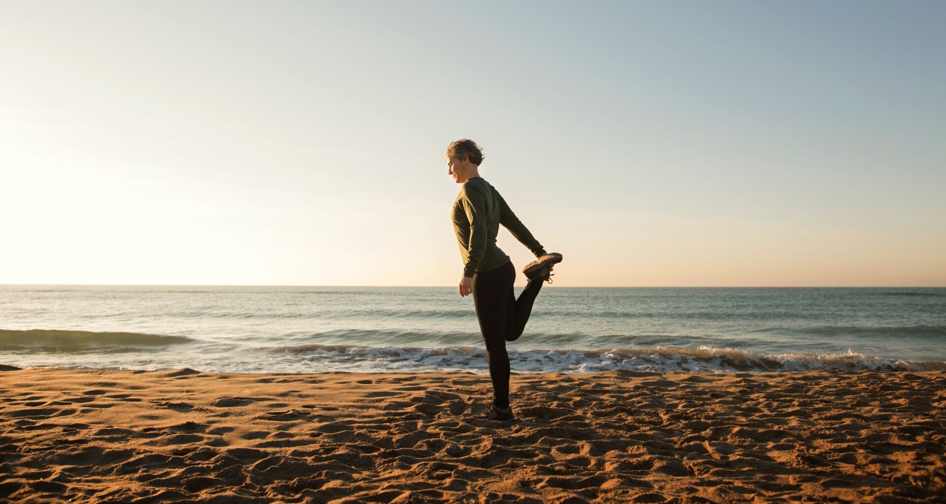 Woman holding one leg standing on the beach next to the sea