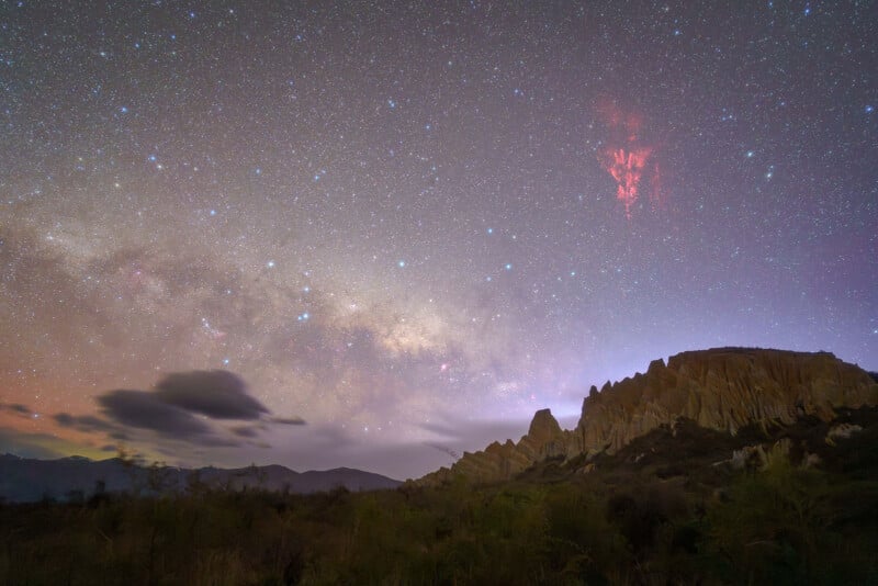A star-filled night sky with a visible section of the Milky Way above rocky cliffs. Wispy clouds and red streaks of atmospheric light appear, with soft landscape silhouettes in the foreground.