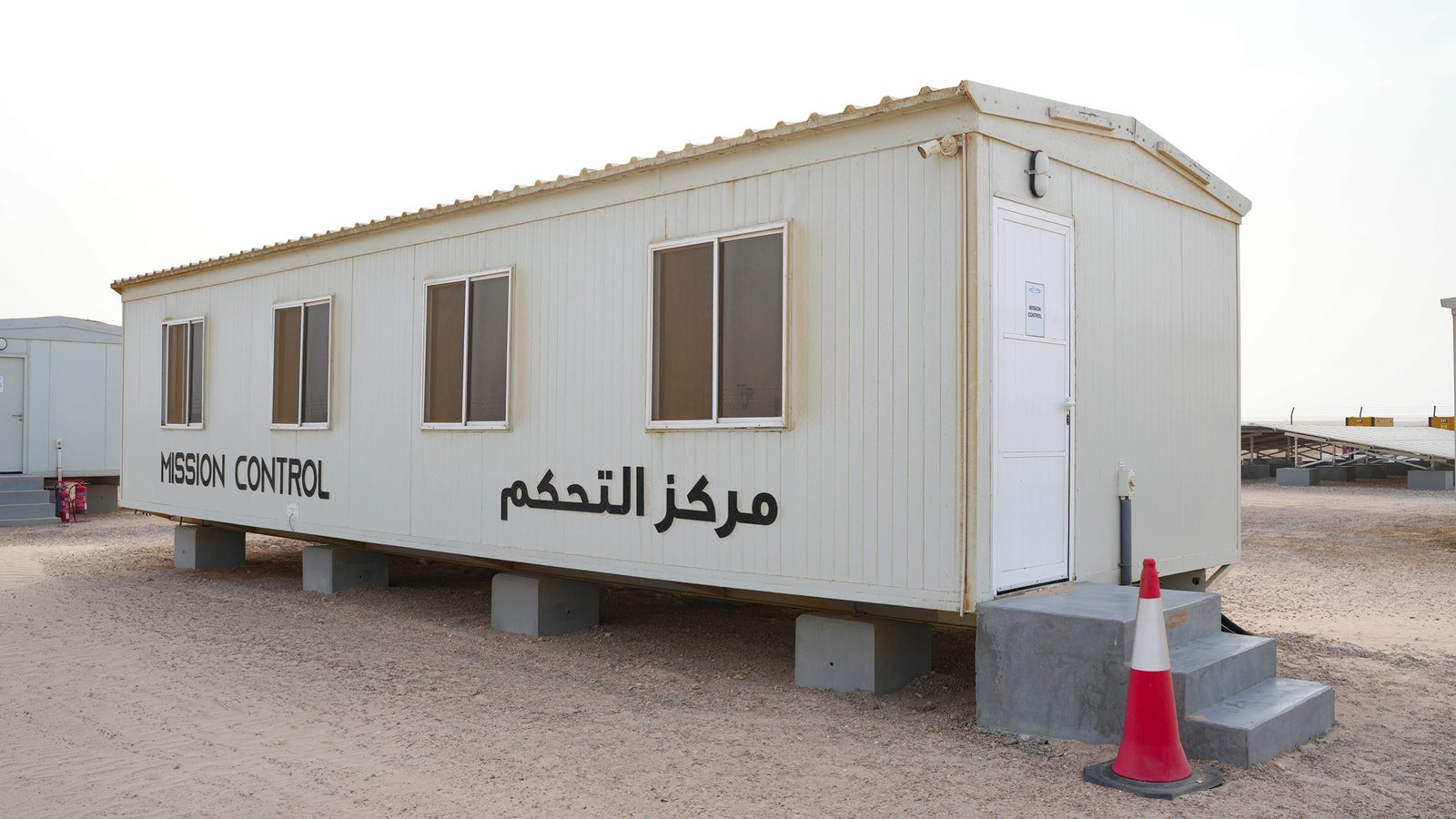 A white modular building labeled 'MISSION CONTROL' in English and Arabic, situated on a sandy surface with several windows and steps leading to the entrance, alongside a red and white traffic cone.