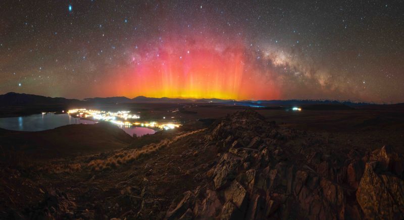 Looking down from a mountain on a town lit up beside the water and in the background are orange and red rays of aurora under an arcing Milky Way.