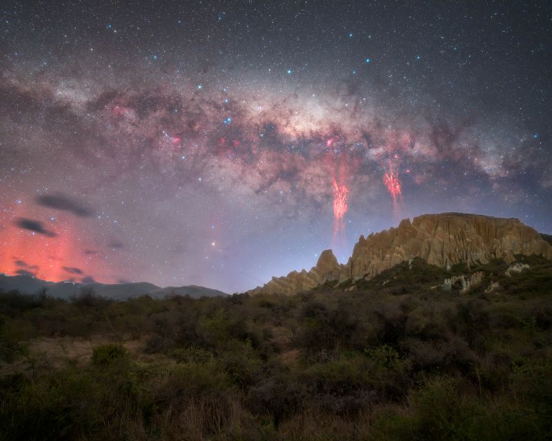 A remote mountainous region with the bright Milky Way and star clouds above, with 2 red sprites in front of a portion of the Milky Way, and a red glow to the left.