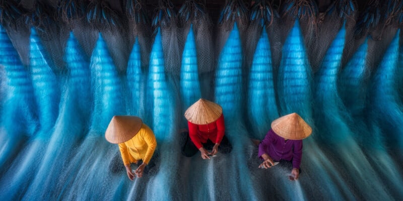 Three people wearing conical hats and colorful clothing sit on blue fishing nets, sorting or weaving, viewed from above, with light streaming through the nets, creating a visually striking pattern.