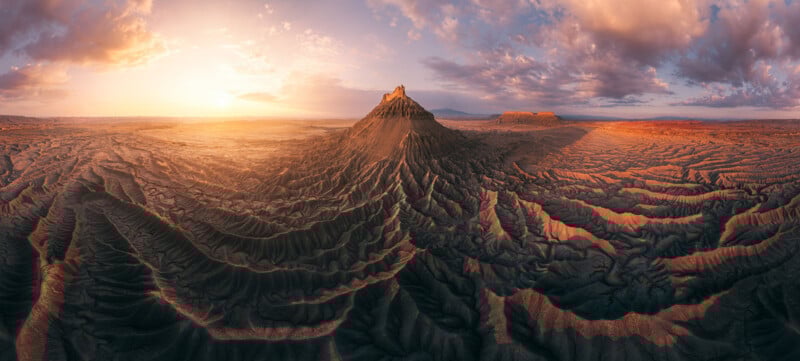 A panoramic view of a rugged desert landscape at sunrise, featuring dramatic ridges, deep shadows, and a central flat-topped mesa under a partly cloudy sky.