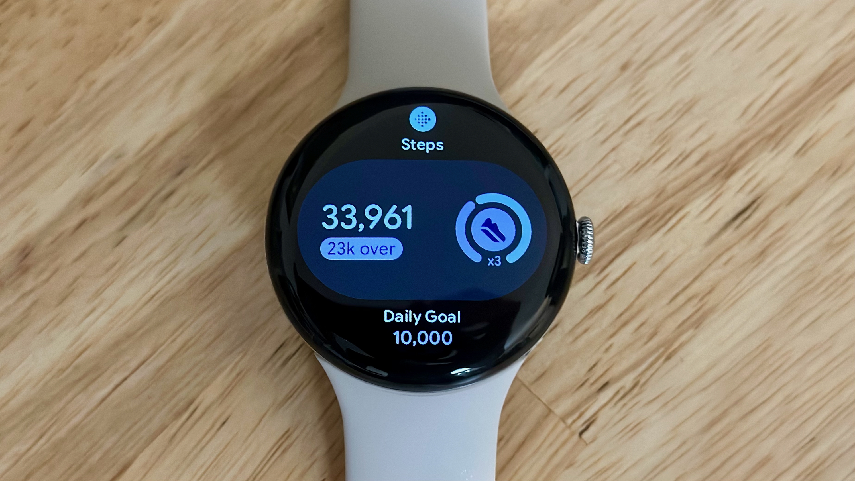 A photo of the Google Pixel Watch 4 on a light wooden desk showing the "Steps" Tile, showing a current count of 33,961 with "23k over" and a Daily Goal of 10,000 underneath.