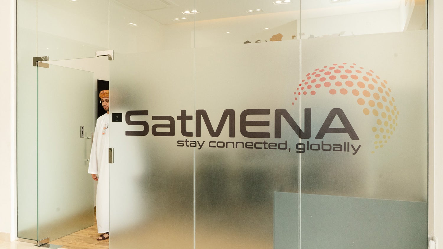 A frosted glass office door with the logo of 'SatMENA' prominently displayed, accompanied by the tagline 'stay connected, globally.' A man dressed in traditional Omani attire stands near the door, partially visible.
