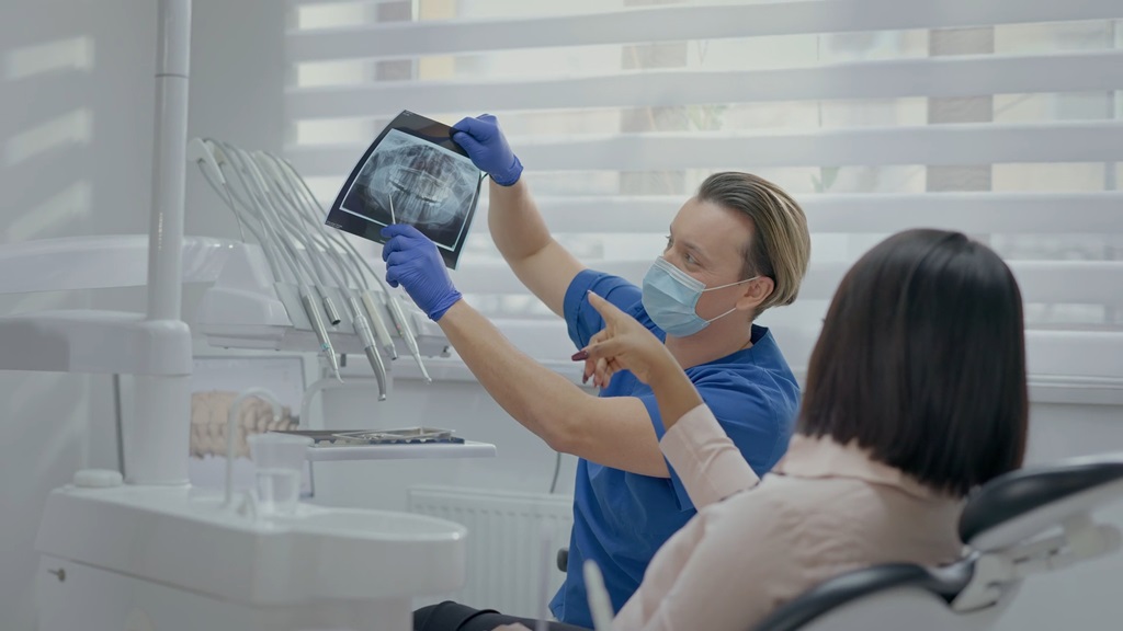 Male orthodontist wearing a medical mask and gloves showing a dental X-ray to a female patient in a modern clinic