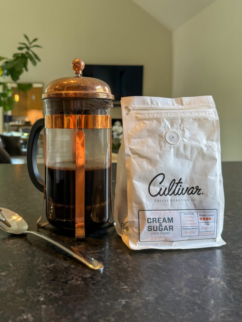 A French press with coffee, a silver spoon, and a bag of Cultivar Cream & Sugar dark roast coffee by Tiffany Woodson sit on a dark countertop in a well-lit kitchen.