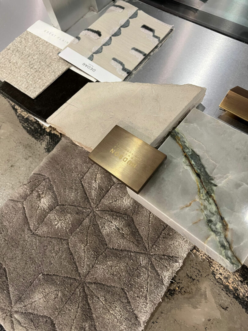 Assorted interior design samples by Tiffany Woodson, including textured fabric, stone, tile, and brass hardware, are artfully arranged on a marble surface.