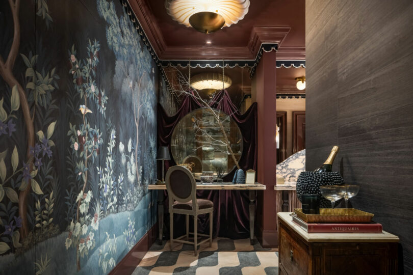 A dimly lit study inspired by Tiffany Woodson, featuring a large round mirror, ornate floral wallpaper, a writing desk with chair, and a side table holding books and a champagne bottle.
