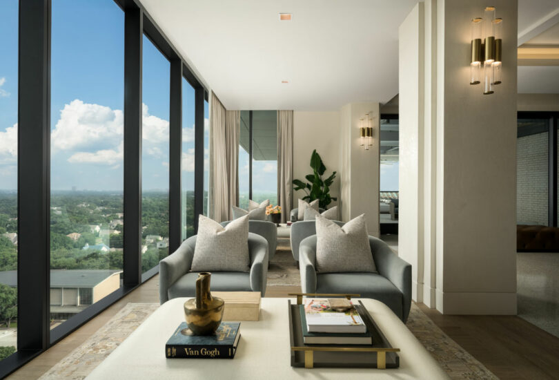 Modern living room designed by Tiffany Woodson, featuring large floor-to-ceiling windows, two gray armchairs, a coffee table with books and decor, and a view of trees and city buildings outside.