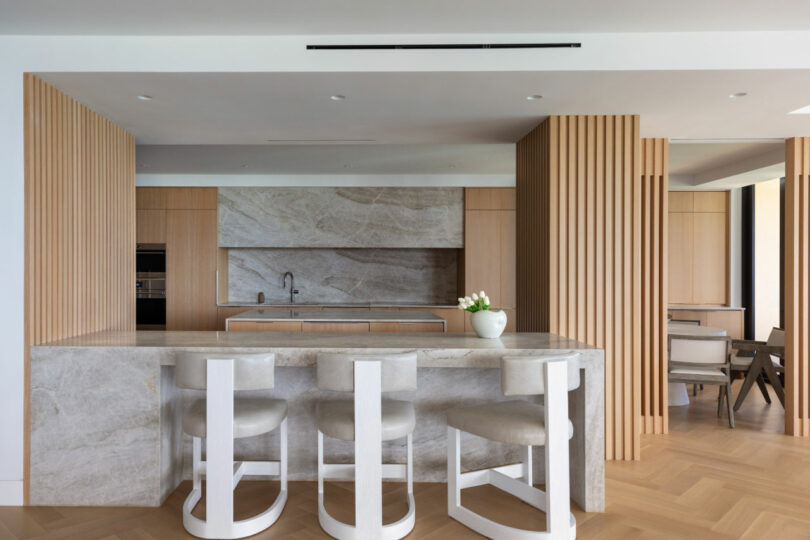Modern kitchen by Tiffany Woodson featuring marble countertops and backsplash, light wood cabinetry, vertical wooden slats, and three white bar stools at the island. A vase with flowers adds a charming touch to the counter.