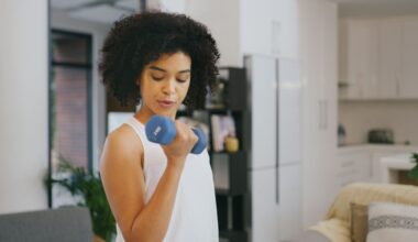 woman curling a blue dumbbell to her biceps, wearing a white vest in a home setting
