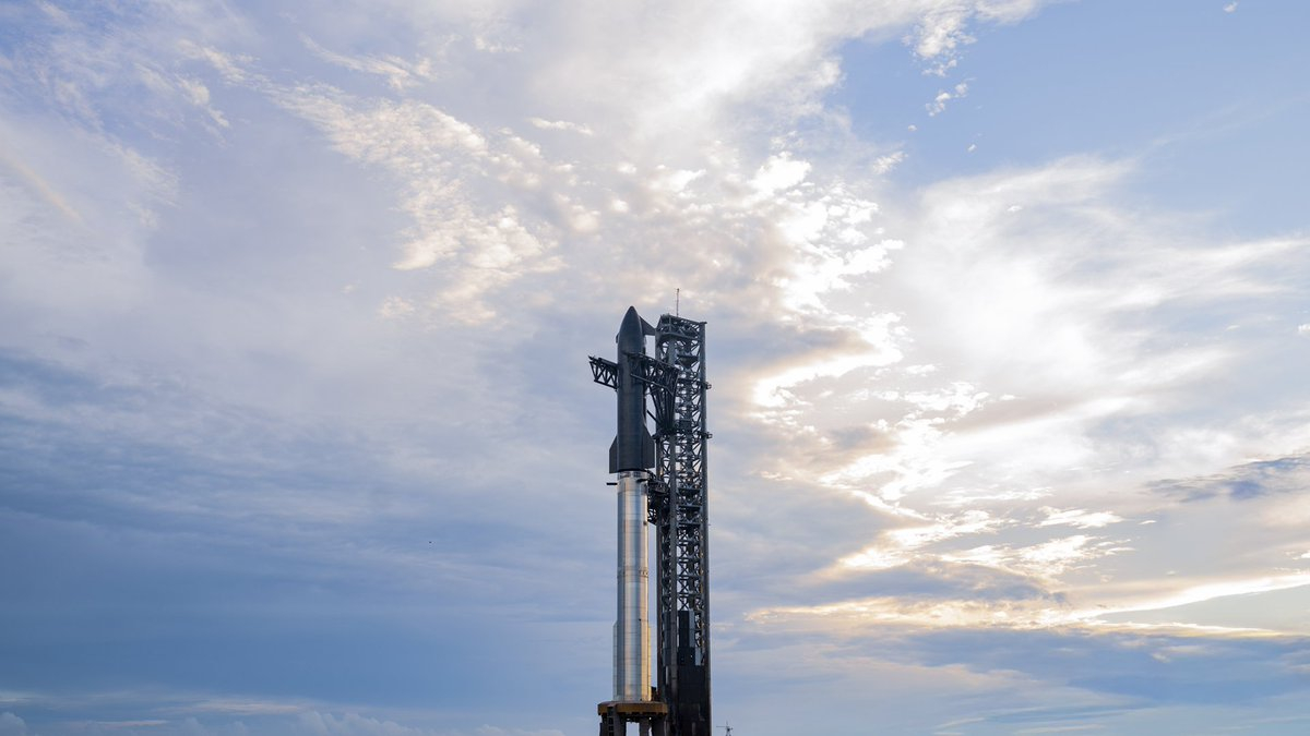 A giant SpaceX Starship rocket on the launch pad with clouds and some blue sky.