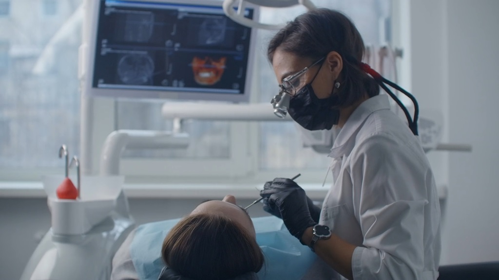 Female orthodontist wearing black gloves and mask examining a patient while viewing 3D dental scans on a nearby monitor in a bright dental clinic