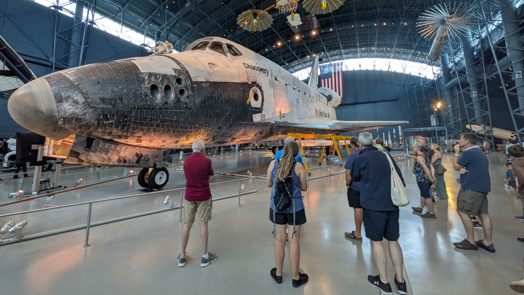 The space shuttle Discovery, a white and black shuttle is on display at the Smithsonian Museum where people walk around it and view it.