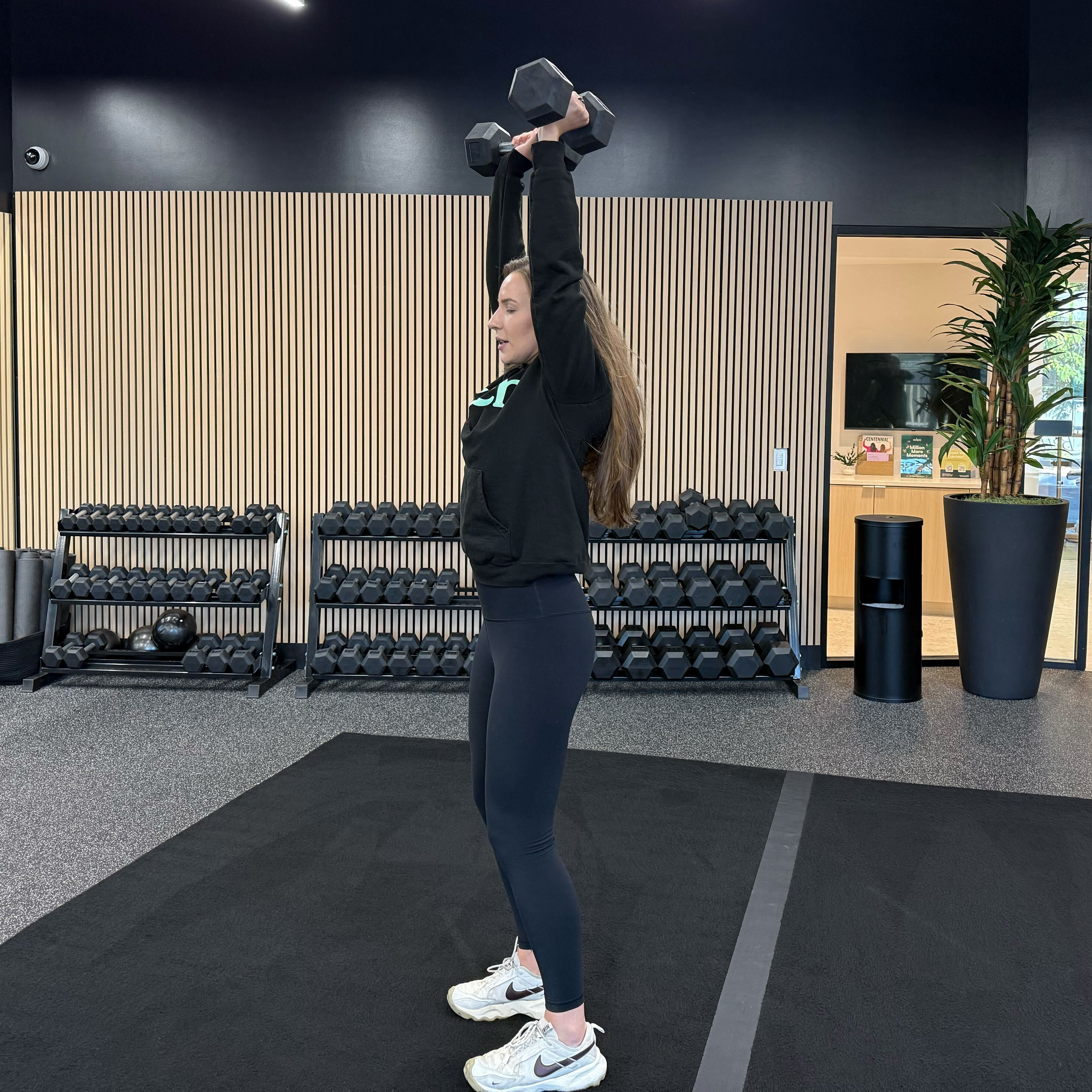 Felicia Hernandez, a personal trainer for Eden Health Club, performs a dumbbell shoulder press in a fitness studio. She is standing with feet shoulder-width apart, arms straight and dumbbells held up toward the ceiling.