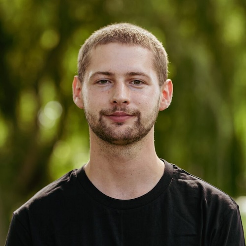 Man with short hair, beard and moustache, wearing a black t-shirt. Blurred trees are behind him.
