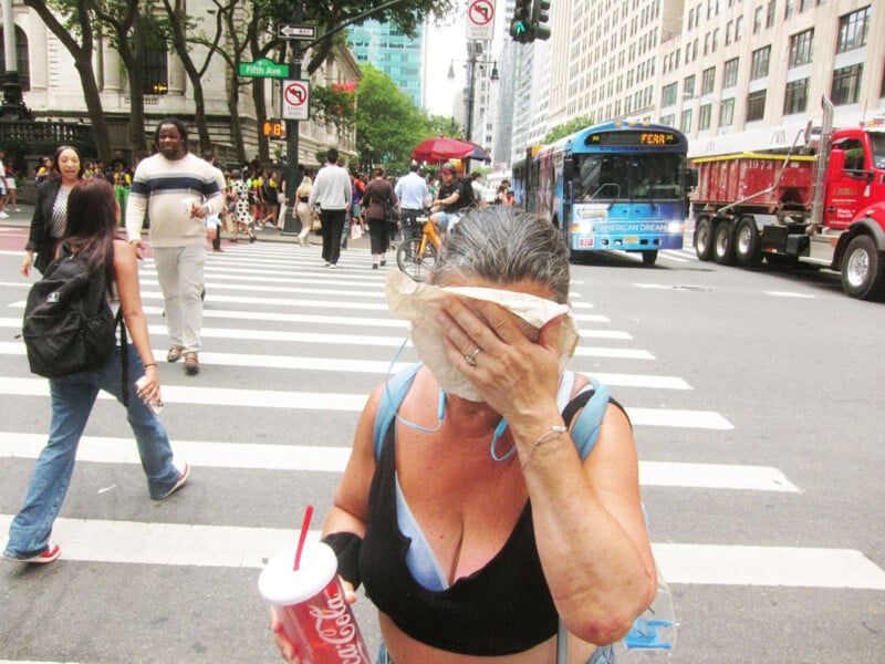 A woman covers her face with a napkin while holding a drink on a busy city crosswalk. People walk nearby, and buses, trucks, and tall buildings line the street in the background.