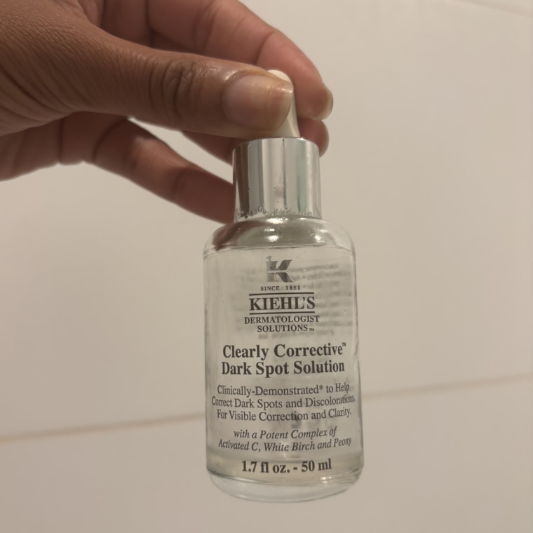 A left hand holding the Kiehl’s Clearly Corrective Dark Spot Corrector in front of a tiled bathroom wall.