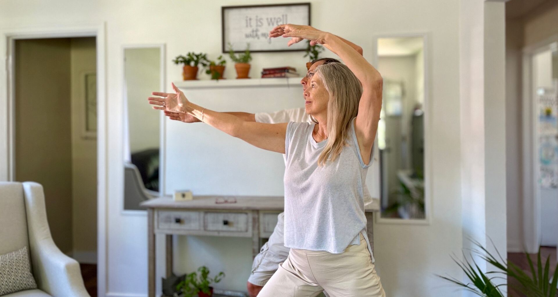 woman standing in front of a man both performing tai chi moves in a living room setting looking sideways away from the camera