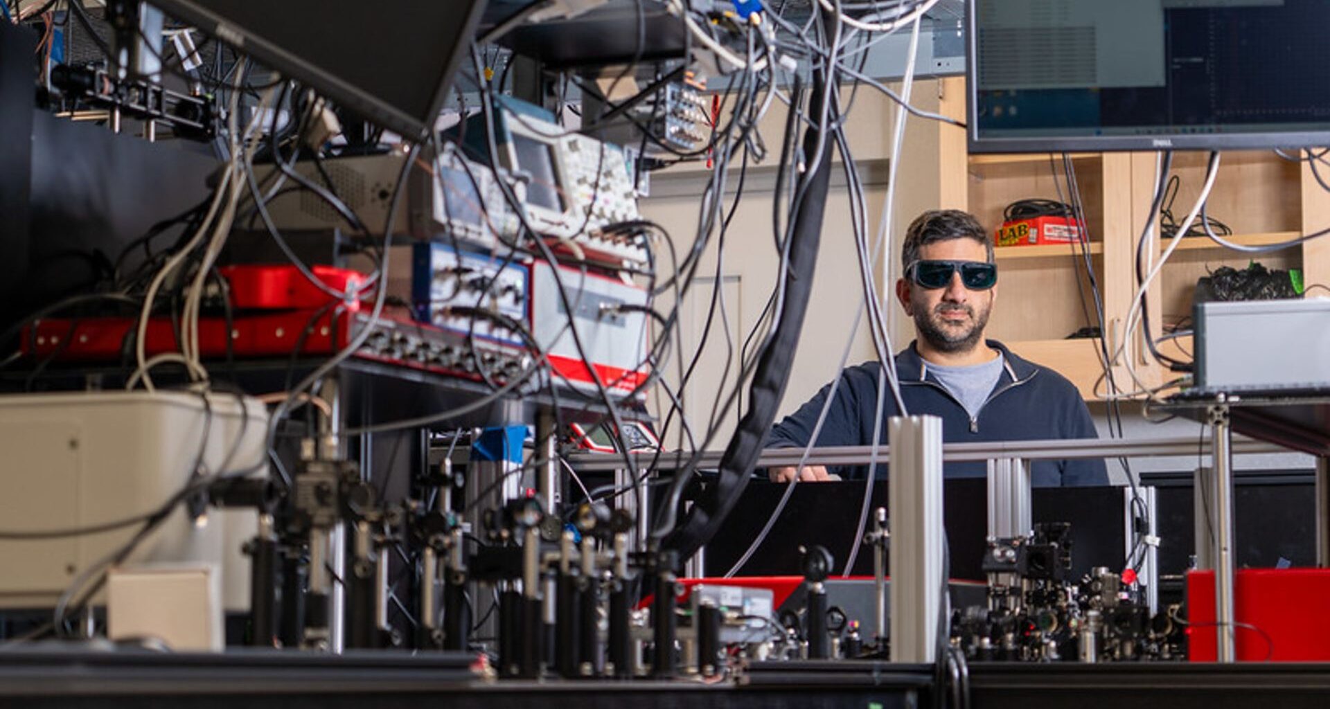 A man wearing dark safety glasses stands behind a table full of wires, mirrors, and lasers