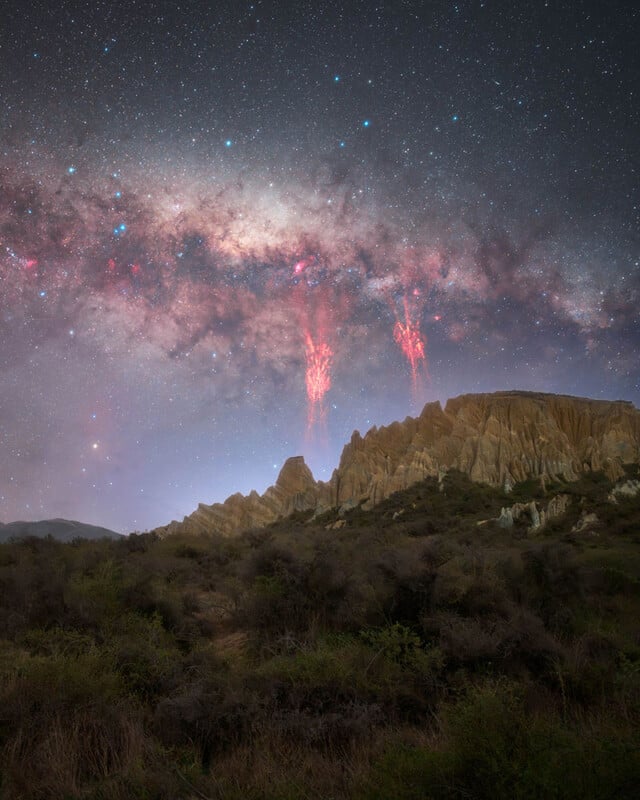 A rugged rocky ridge rises above brushy terrain under a star-filled night sky, with the Milky Way and two vivid red lightning-like sprites illuminating the atmosphere above the ridge.